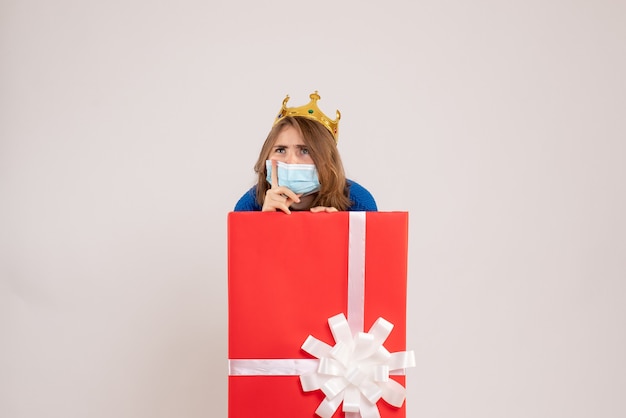 Vorderansicht der jungen Frau in Geschenkbox in Maske auf weißer Wand