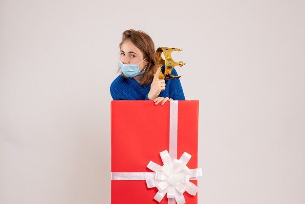 Vorderansicht der jungen Frau in Geschenkbox in Maske auf weißer Wand