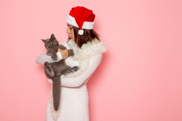 Vorderansicht der jungen Frau in roter Mütze, die ein süßes graues Kätzchen an der rosa Wand hält