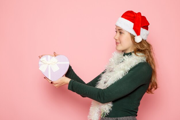 Vorderansicht der jungen Frau in roter Mütze, die herzförmiges Geschenk an rosa Wand hält