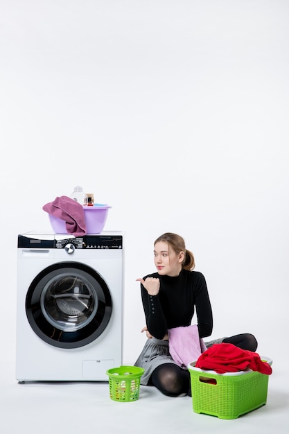 Vorderansicht der jungen Frau mit Waschmaschine, die schmutzige Kleidung auf weißer Wand faltet