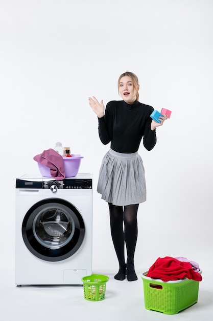 Vorderansicht der jungen Frau mit Waschmaschine und schmutziger Kleidung auf weißer Wand