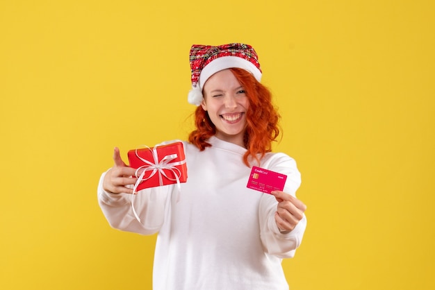 Vorderansicht der jungen Frau mit Weihnachtsgeschenk und Bankkarte auf gelber Wand