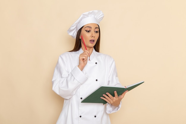 Vorderansicht der jungen Köchin im weißen Kochanzug, der Heft hält und an hellweiße Wand denkt