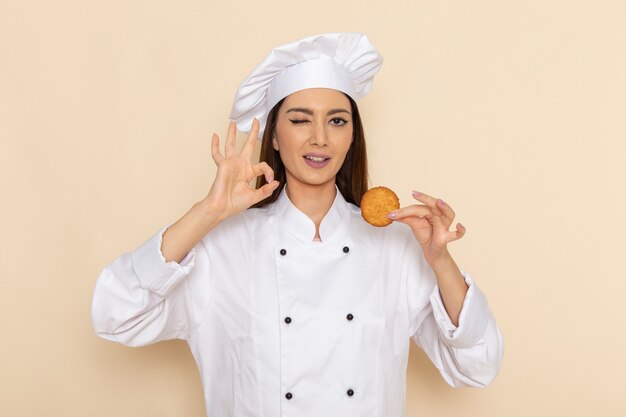 Vorderansicht der jungen Köchin im weißen Kochanzug, der Kekse auf hellweißer Wand hält