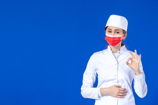 Vorderansicht der jungen Krankenschwester im medizinischen Anzug mit roter Maske auf Blau