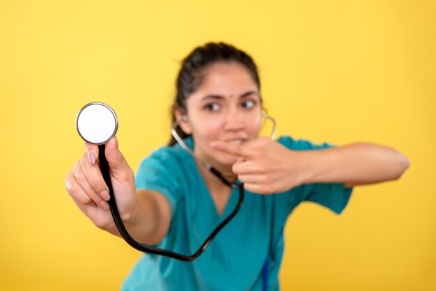 Vorderansicht der jungen Ärztin, die auf Stethoskop auf gelber Wand zeigt