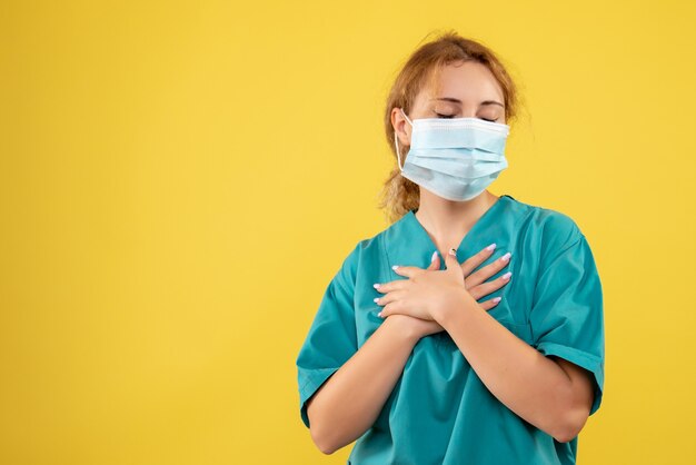 Vorderansicht der jungen Ärztin im medizinischen Anzug und in der sterilen Maske auf gelber Wand