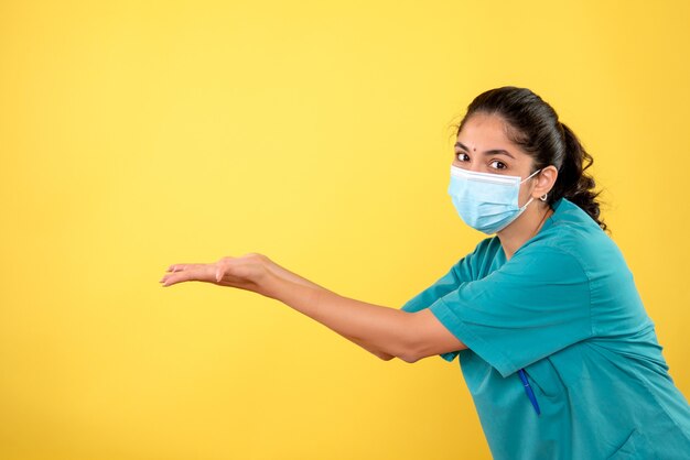 Vorderansicht der jungen Ärztin mit medizinischer Maske, die ihre Hände auf gelber Wand öffnet