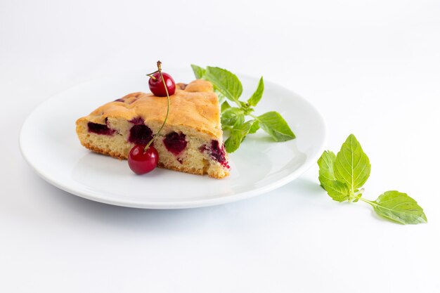 Vorderansicht der Kirschkuchenscheibe innerhalb der weißen Platte mit grünen Baumblättern auf der weißen Oberfläche