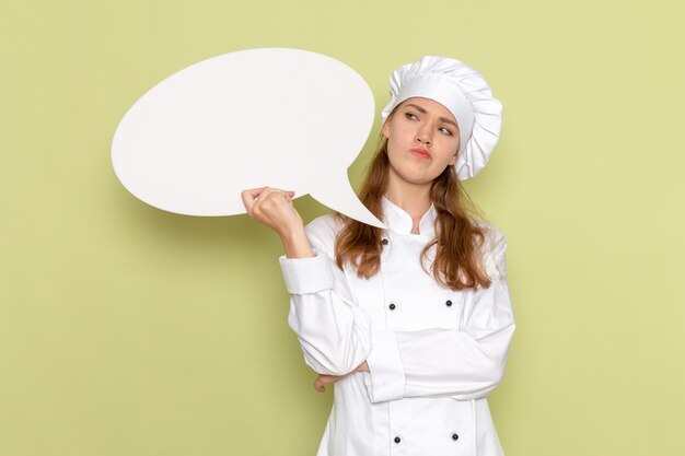 Vorderansicht der Köchin, die weißen Kochanzug hält, weißes Zeichen hält und auf hellgrüne Wand denkt