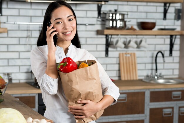 Vorderansicht der lächelnden asiatischen Frau, die am Handy beim Halten der Einkaufstüte spricht