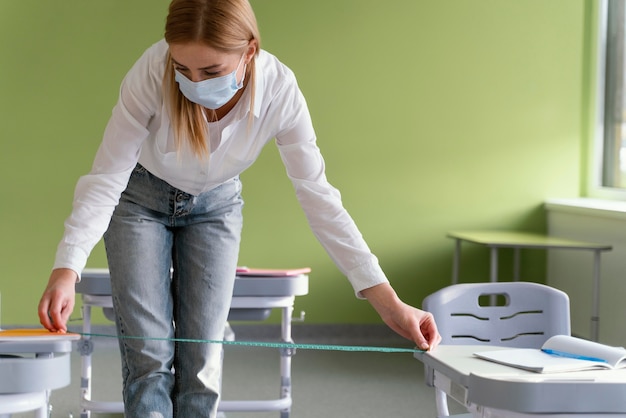 Vorderansicht der Lehrerin mit medizinischer Maske, die den Abstand zwischen Klassenbänken misst