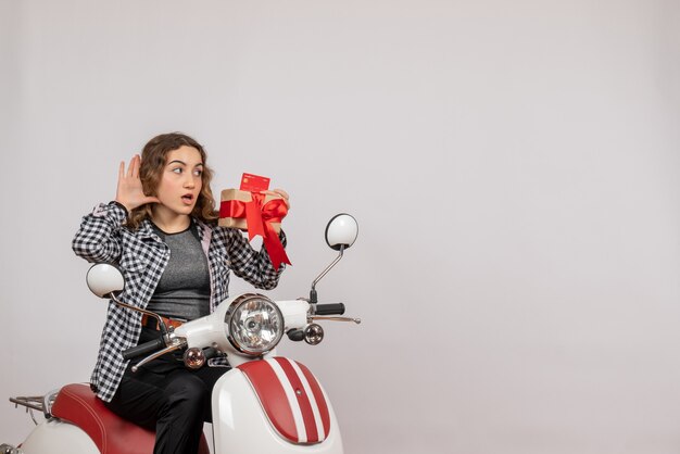 Vorderansicht der neugierigen jungen Frau auf Moped, das Geschenk hält, das etwas auf grauer Wand hört