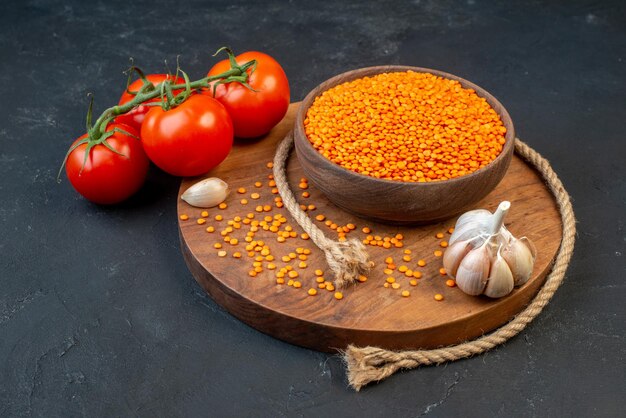 Vorderansicht der roten Linsen in einer braunen Schüssel Seilknoblauch auf hölzernen runden Bretttomaten mit Stielen auf der rechten Seite auf schwarzem Hintergrund