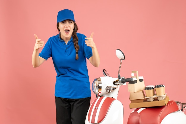 Vorderansicht der sich wundernden Kurierdame, die neben dem Motorrad mit Kaffee und kleinen Kuchen auf pastellfarbenem Hintergrund steht