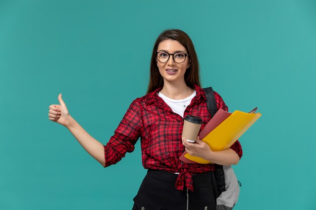 Vorderansicht der Studentin, die Rucksack hält, der Dateien und Kaffee auf hellblauer Wand hält