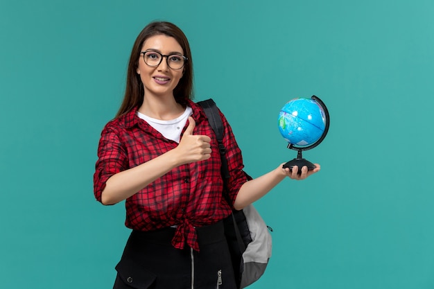 Vorderansicht der Studentin, die Rucksack hält, der kleinen Globus auf hellblauer Wand hält