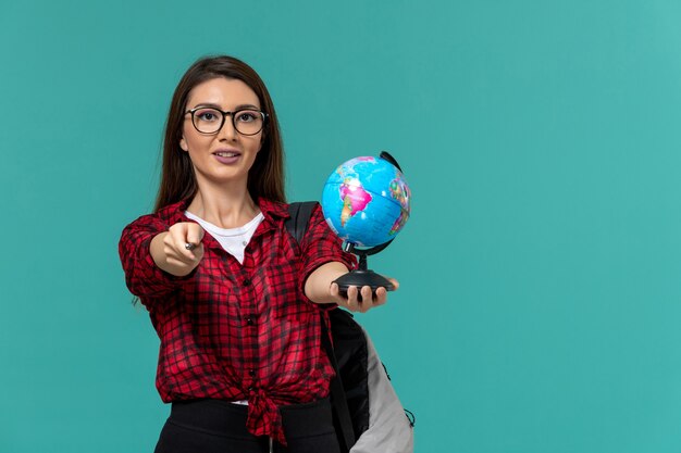 Vorderansicht der Studentin, die Rucksack hält, der kleinen Globus und Stift auf hellblauer Wand hält