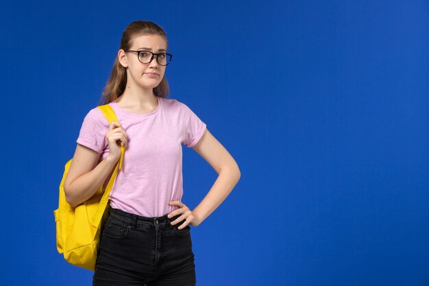 Vorderansicht der Studentin im rosa T-Shirt mit gelbem Rucksack an der hellblauen Wand