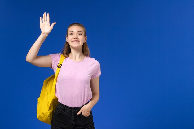 Vorderansicht der Studentin im rosa T-Shirt mit gelbem Rucksack, der auf der blauen Wand aufwirft und lächelt