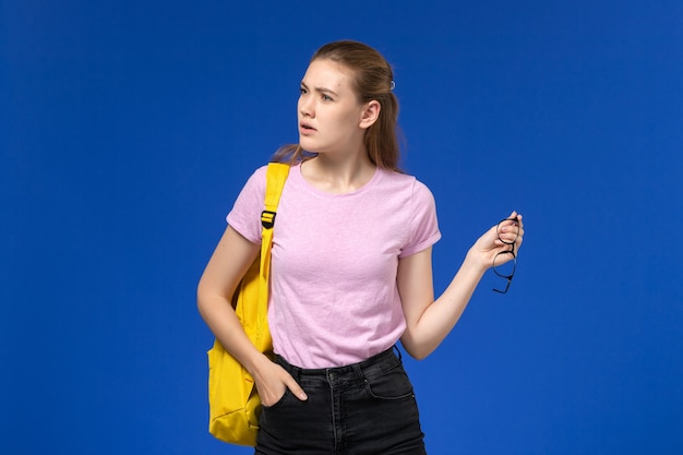 Vorderansicht der Studentin im rosa T-Shirt mit gelbem Rucksack, der auf der blauen Wand aufwirft