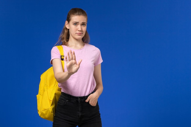 Vorderansicht der Studentin im rosa T-Shirt mit gelbem Rucksack, der auf der hellblauen Wand aufwirft