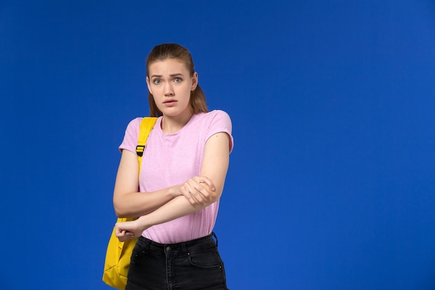 Vorderansicht der Studentin im rosa T-Shirt mit gelbem Rucksack, der gerade auf der blauen Wand steht