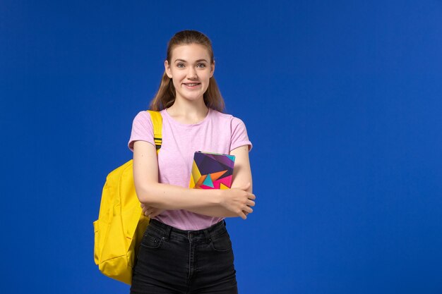 Vorderansicht der Studentin im rosa T-Shirt mit gelbem Rucksack, der Heft an der blauen Wand hält