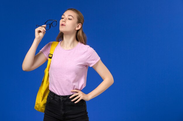 Vorderansicht der Studentin im rosa T-Shirt mit gelbem Rucksack, der optische Sonnenbrille auf hellblauer Wand hält