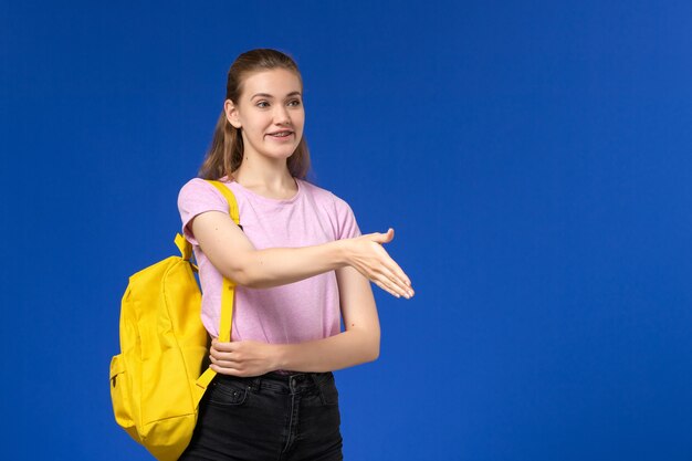 Vorderansicht der Studentin im rosa T-Shirt mit gelbem Rucksack lächelnd und Händeschütteln auf blauer Wand