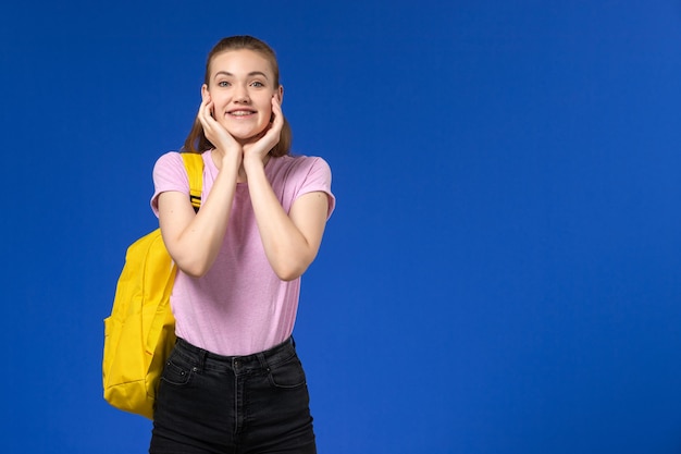 Vorderansicht der Studentin im rosa T-Shirt mit gelbem Rucksack lächelnd und posierend auf der blauen Wand