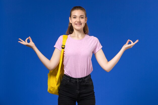 Vorderansicht der Studentin im rosa T-Shirt mit gelbem Rucksack lächelnd und posierend auf der blauen Wand