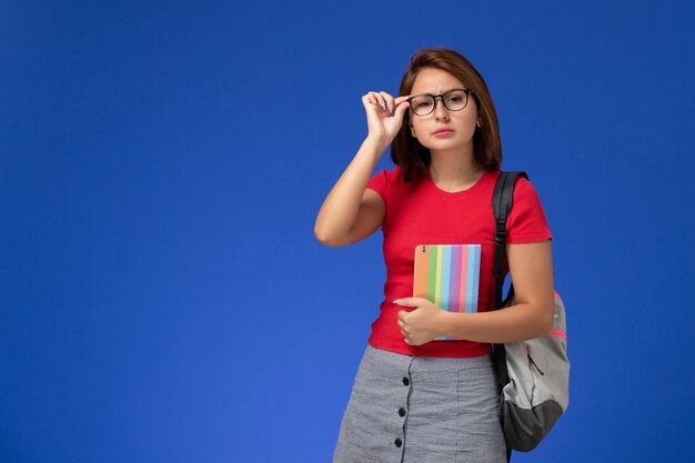 Vorderansicht der Studentin im roten Hemd mit dem Rucksack, der das Heft an der blauen Wand hält