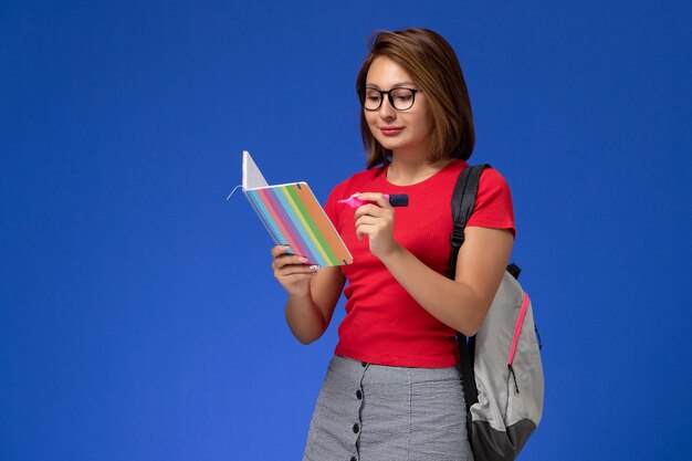 Vorderansicht der Studentin im roten Hemd mit dem Rucksack, der Filzstifte hält, die Heft auf der blauen Wand lesen