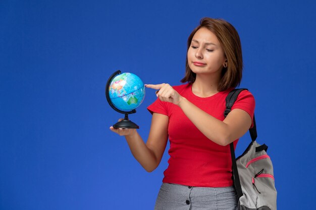 Vorderansicht der Studentin im roten Hemd mit dem Rucksack, der kleinen Globus auf der blauen Wand hält