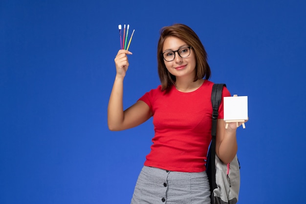 Vorderansicht der Studentin im roten Hemd mit dem Rucksack, der Quasten hält, zum Lächeln lächelnd auf der hellblauen Wand