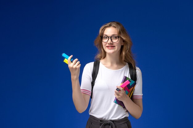 Vorderansicht der Studentin im weißen Hemd, das Rucksack trägt und Heft mit Filzstiften an der blauen Wand hält