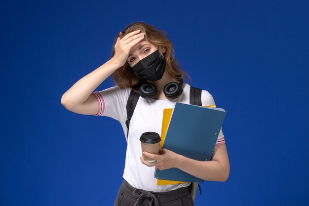 Vorderansicht der Studentin im weißen Hemd, das schwarze sterile Rucksackmaske hält, die Kaffee und Dateien an der blauen Wand hält