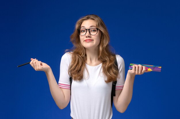 Vorderansicht der Studentin im weißen Hemd, das Stift und Heft an der blauen Wand hält