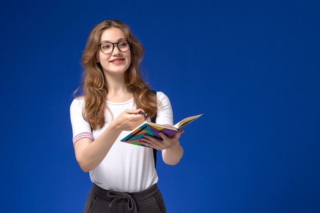 Vorderansicht der Studentin im weißen Hemd, das Stift und Heft mit Lächeln auf der blauen Wand hält