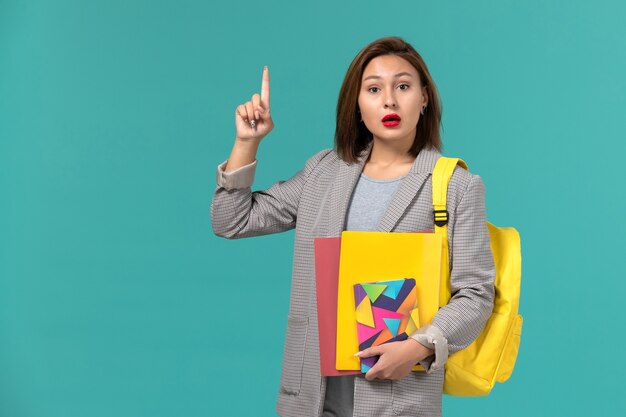 Vorderansicht der Studentin in der grauen Jacke, die gelben Rucksack hält, der Dateien und Heft an der hellblauen Wand hält