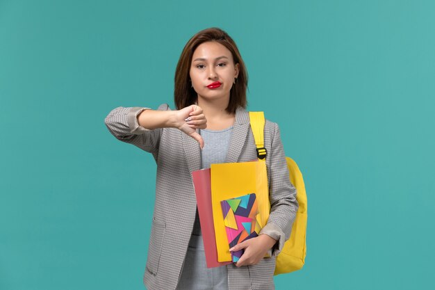 Vorderansicht der Studentin in der grauen Jacke, die gelben Rucksack hält, der Dateien und Heft hält, das unähnliches Zeichen auf blauer Wand zeigt