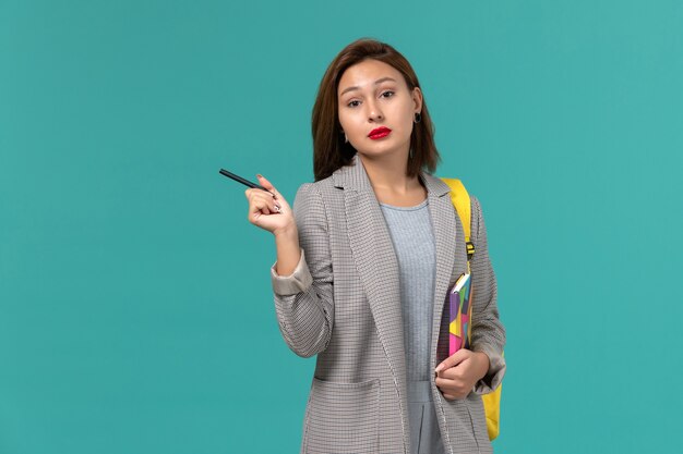 Vorderansicht der Studentin in der grauen Jacke, die gelben Rucksack hält, der Heft mit Stift auf der blauen Wand hält