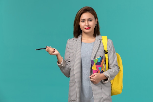 Vorderansicht der Studentin in der grauen Jacke, die gelben Rucksack hält, der Heft mit Stift auf der blauen Wand hält