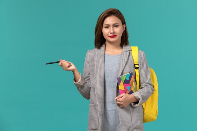 Vorderansicht der Studentin in der grauen Jacke, die gelben Rucksack hält, der Heft mit Stift auf der blauen Wand hält