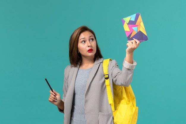 Vorderansicht der Studentin in der grauen Jacke, die gelben Rucksack hält, der Heft mit Stift auf der hellblauen Wand hält