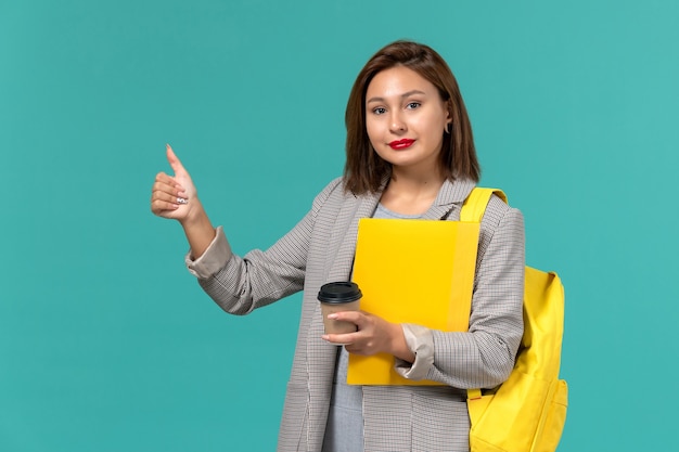 Vorderansicht der Studentin in der grauen Jacke, die ihren gelben Rucksack hält, der Dateien und Kaffee auf der hellblauen Wand hält