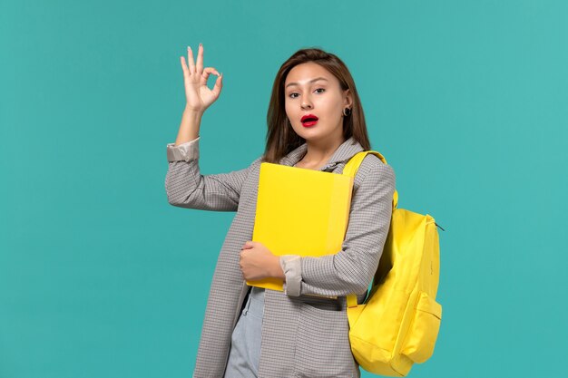 Vorderansicht der Studentin in der grauen Jacke, die ihren gelben Rucksack trägt und Akten an der hellblauen Wand hält