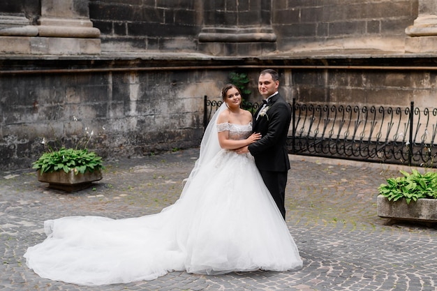 Vorderansicht der süßen Braut im Hochzeitskleid mit langer Schleppe und Schleier, die in der Nähe des Bräutigams steht und seine Hand hält, die während des Hochzeitsspaziergangs in der antiken Stadt in die Kamera blickt und posiert